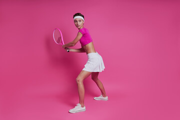 Attractive young woman holding tennis racket while standing against pink background