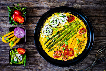 Tasty breakfast. Omelette  with green asparagus, goat cheese, pepper and cherry tomatoes served on black plate on wooden table
