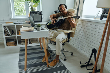 Confident young African man playing acoustic guitar while sitting at his working place