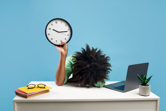 Tired black male student lying at desk with pc laptop and books, holding clock and putting head on hand, blue background