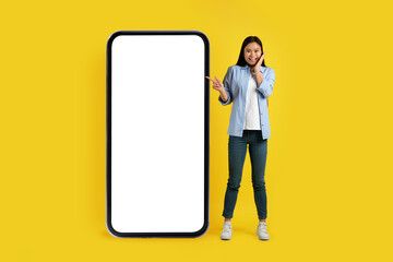 Happy surprised young chinese female pointing at huge phone with empty screen, isolated on yellow background