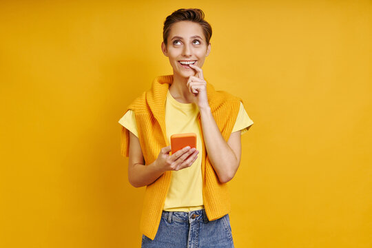 Dreamful young woman holding smart phone while standing against yellow background