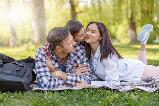 Happy family with kid lying on green grass in park. Mother and father hugging and kissing son.