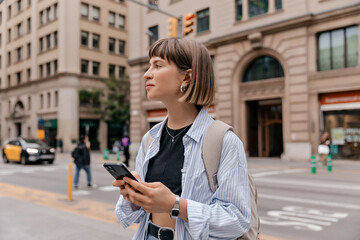 Spectacular happy caucasian girl with short hair wearing blue shirt with backpack is holding smartphone and looking aside on city background in warm day