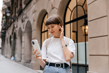 Beautiful young caucasian woman walks near hotel building. Portrait of successful stylish woman holding smartphone and touching headphones in her hand on way to work on city street.