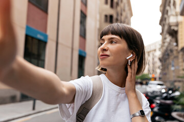 Romantic lovely girl with short straight hair in headphones is making selfie and looking aside on city background. Closeup selfie-portrait student of attractive girl in city.