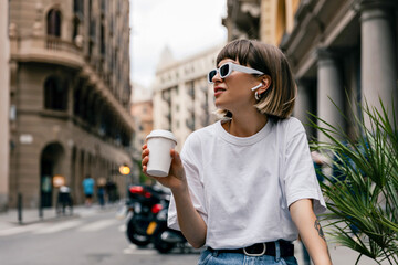 Stylish european girl with short hair wearing shite t-shirt in sunglasses drinking coffee an...