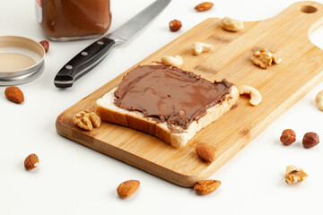 Square bread for toast with chocolate spread on a wooden board. Nuts, a knife, a jar of chocolate paste, and a wooden cutting board with a sandwich on a white table.
