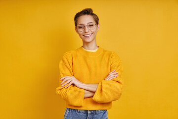 Beautiful young woman keeping arms crossed while standing against yellow background