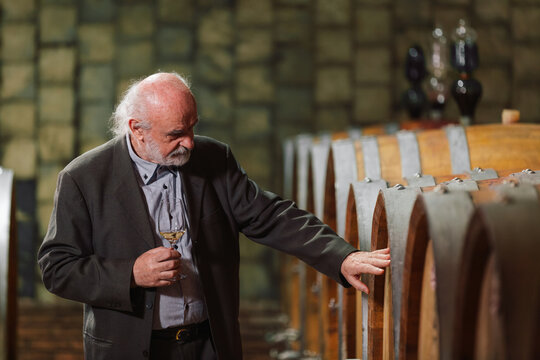 Senior Caucasian graybeard man enjoying visiting wine cellar, looking around wooden barrels and tasting red wine, showing pleasure and contentment