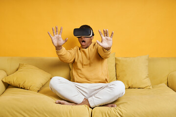 Shocked young man in virtual reality glasses relaxing on the couch with yellow background