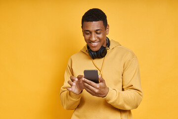 Handsome African man with headphones using smart phone while standing against yellow
