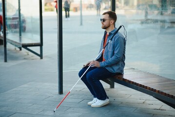 Visually impaired man with walking stick, sitting on bench in city park. Copy space