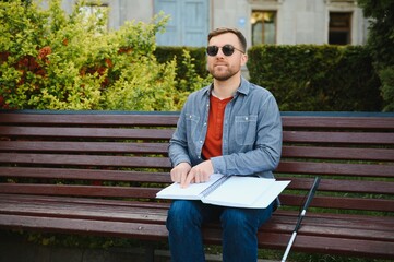 Blind man reading braille book, sitting on bench in summer park, resting