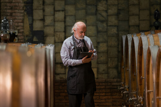 Caucasian senior man, a graybeard winemaker with apron checking big wooden barrels in a wine cellar and entering data in a tablet device