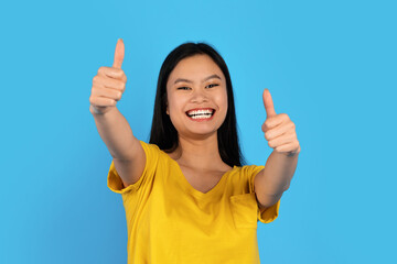 Portrait of happy young asian lady with down syndrome in yellow t-shirt showing thumbs up on blue background