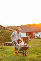 Happy little children having fun in a wheelbarrow pushing by dad in domestic garden on warm sunny day. Active outdoors games for family with kids. Spending time together on weekend at the countryside.