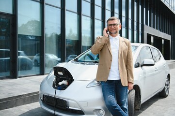 The guy sat down on the hood of the car. His car is charging at the charging station. A man looks...