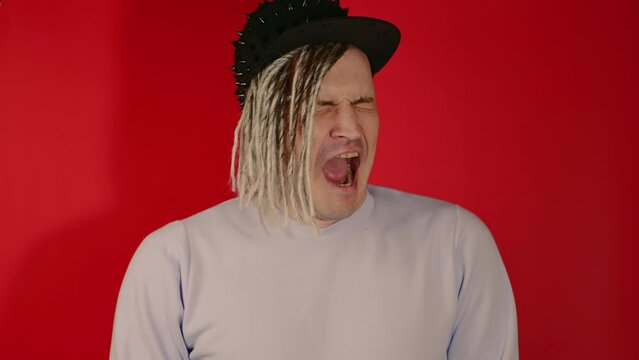 Young tired handsome man in black cap yawning, closing eyes, opening mouth. Exhausted guy with blonde dreadlocks feeling fatigue, yawning, covering mouth with hand on red background in studio.