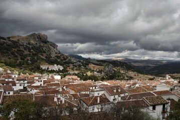 Obraz premium Red terracotta roofs of the white village of Grazalema, Spain, against a backdrop of spectacular dark storm clouds and the dry autumn slopes of yellow brown grass of the Sierra del Pinar mountains