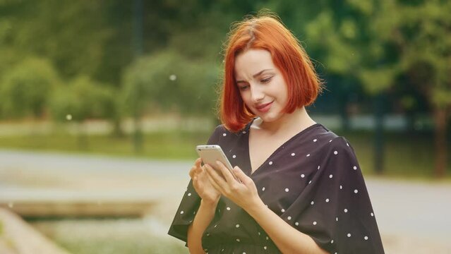 Beautiful focused redhead caucasian girl teenage user woman with short hair stand in city park hold modern device smartphone gadget look at mobile screen read chatting message use urban wi-fi internet