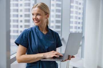 Smiling female healthcare worker using laptop while working at doctor's office and looking window