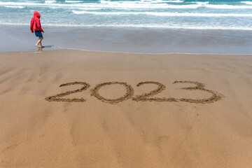 Year 2023 written in the sand on the beach with a boy in a red coat and the sea in the background.