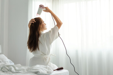 Young Asian woman in bathrobe uses hair dryer on bed.