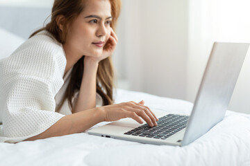 Pretty Asian woman lying down the bed while using laptop computer at home bedroom, close up, selective focus