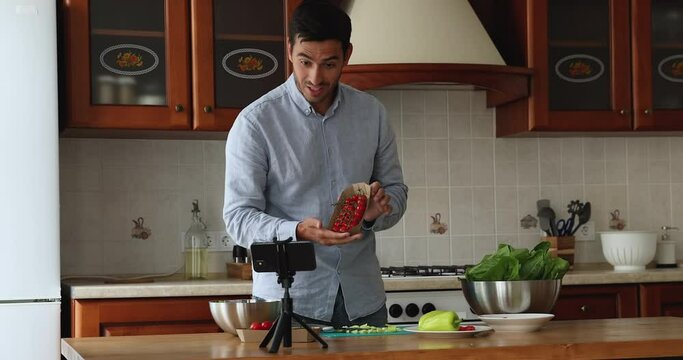 Happy handsome cook blogger man recording video on smartphone in kitchen, giving workshop, preparing salad, showing fresh vegetable ingredients at camera on tripod stand. Eating, marketing concept