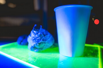 White paper cup with coffee and delicious cake and cookies over a neon green tray in a coffee shop interior in the night with dark blue lights