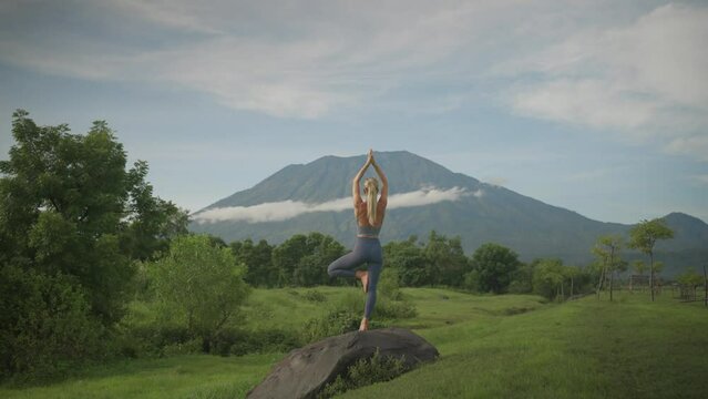 Athletic blond woman in yoga tree pose balancing on rock, Mount Agung in background