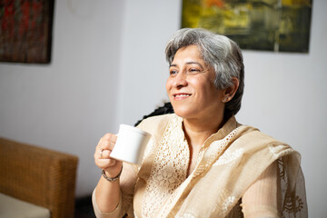 Aging, Happy indian senior woman sitting in living room with cup of tea or coffee, looking away, Retired middle aged lady enjoying no stress calm positive pastime alone at home.