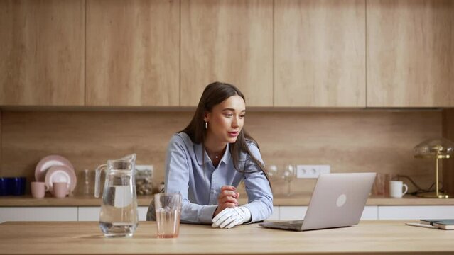 Young disabled woman is chatting in front of laptop at table in home kitchen spbas. 4k Close view of attractive female with prosthetic arm looks at screen and speaks with smile, discusses something