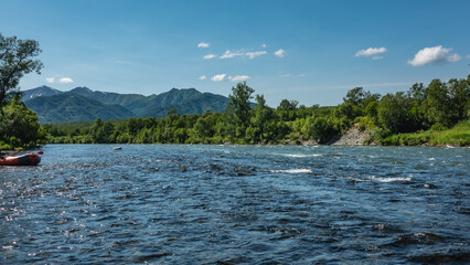 Ripples and foam are visible on the surface of the mountain river. An inflatable rafting boat is moored near the shore. Green forest and mountains against the blue sky. Kamchatka. River Bystraya