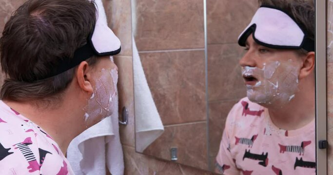Tired man with bruises under eyes wearing sleep mask on head stands with foam on face looks in mirror , sighs discontentedly. Hangover after bachelor party, consequences of sleepless night.