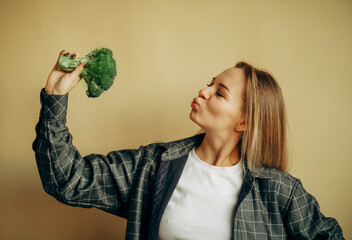 Photo of cheerful young blond woman standing over beige background while holding broccoli