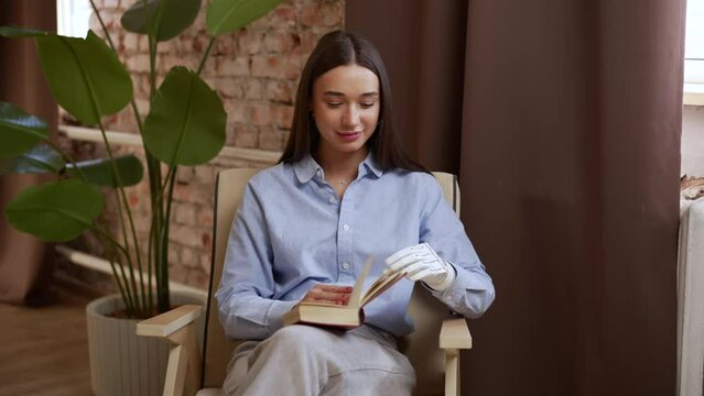 Young disabled woman is reading book and smiling while sitting in chair in home interior spbas. 4k Close view of European female with bionic hand opens book and looks with smile, poses for camera and