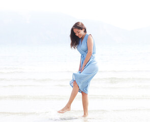 Fototapeta na wymiar Young brunette woman in a blue dress walking barefoot on a beach and dangles his feet in the water.