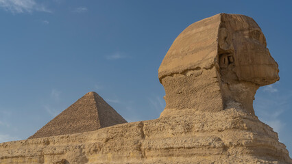 Sculpture of the Great Sphinx. Close-up. Profile view. The head and part of the back are visible. Layered structure of a stone statue. The top of the pyramid against a clear blue sky. Egypt. Giza