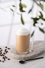 Cappuccino or latte with milk foam in a cup with coffee beans and spoon  on light background with branch and morning shadow