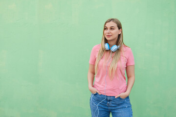 Beautiful cheerful blonde woman with blue headphones in a pink t-shirt stands outdoors against a green wall