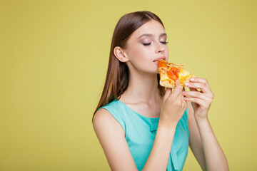 Beautiful young happy woman in a pretty aqua blue dress with  posing on yellow  background. Slim figure, studio shot. Delicious slice of pizza in hands. Model eats pizza