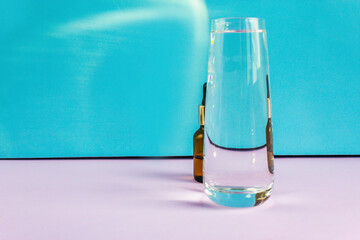 A glass of water and a cosmetic bottle with a pipette on a purple and blue background. Distort the picture through glass and water
