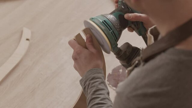 Over the shoulder shot of carpenter using angle grinder while sanding and polishing carved piece of wood, making furniture in workshop