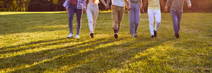 Legs of several men and women holding hands walking along green grass towards camera symbolizing joint walks in friendly company or team building for startup in summer park. Panoramic shot