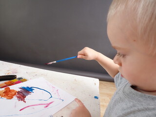 The child learns to draw with a brush with multicolored acrylic paints and gouache on a white sheet...