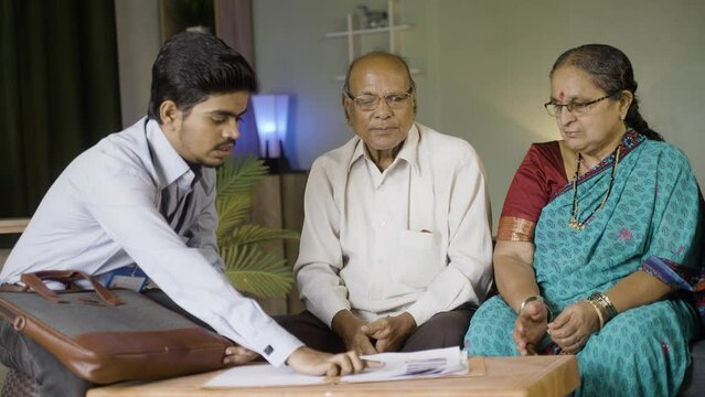Banker advising about insurance to elderly couple while sitting on sofa at home - concept of banking, financial and communication.