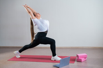 Young Plus Size Woman Stretching At Home Online. Flexible girl practices yoga and watches an online course on a laptop