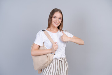 A beautiful girl in white t-shirt with backpack on her back, student smiles cutely on a gray background in the studio, a happy young woman rejoices at the day off. Advertising and mockup